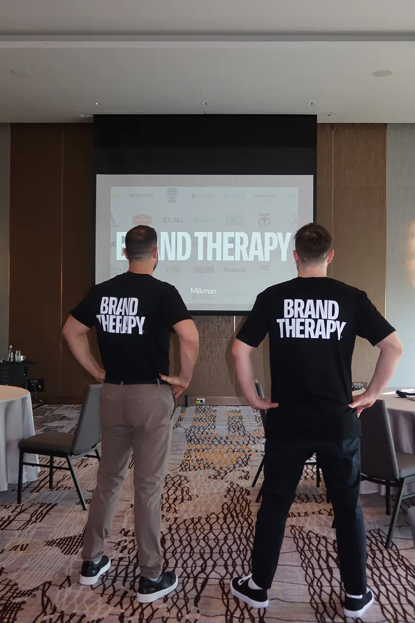 Two facilitators in Brand Therapy T-shirts facing a screen of client logos